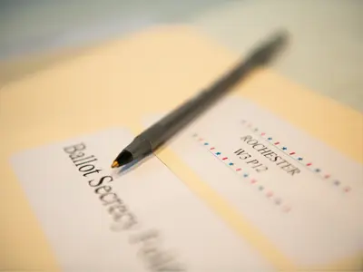 an out of focus ballot secrecy folder that reads 'rochester w3 p12' and has a pen resting on it