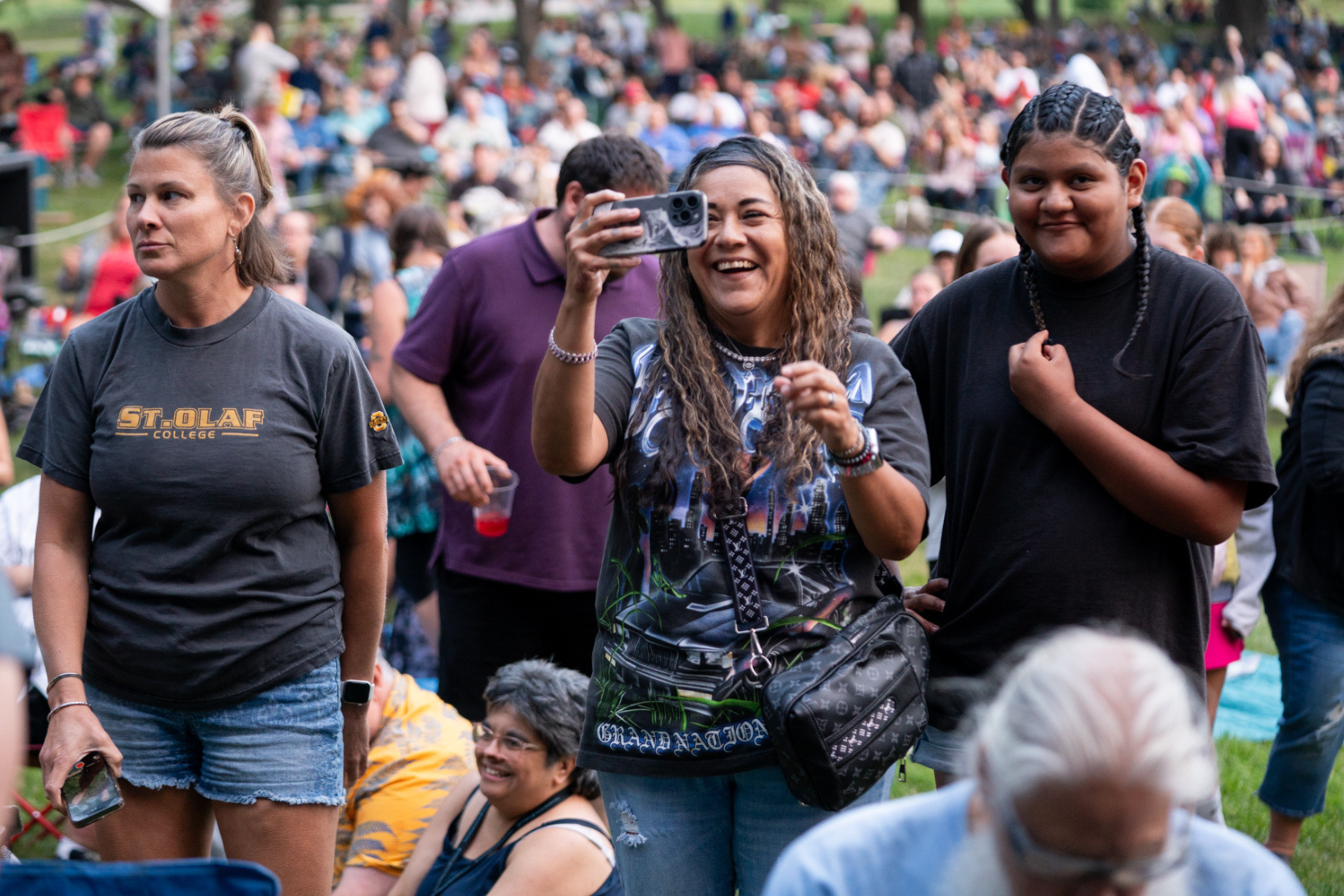 Three people in focus standing in a crowd smiling at something off screen and holding up a phone
