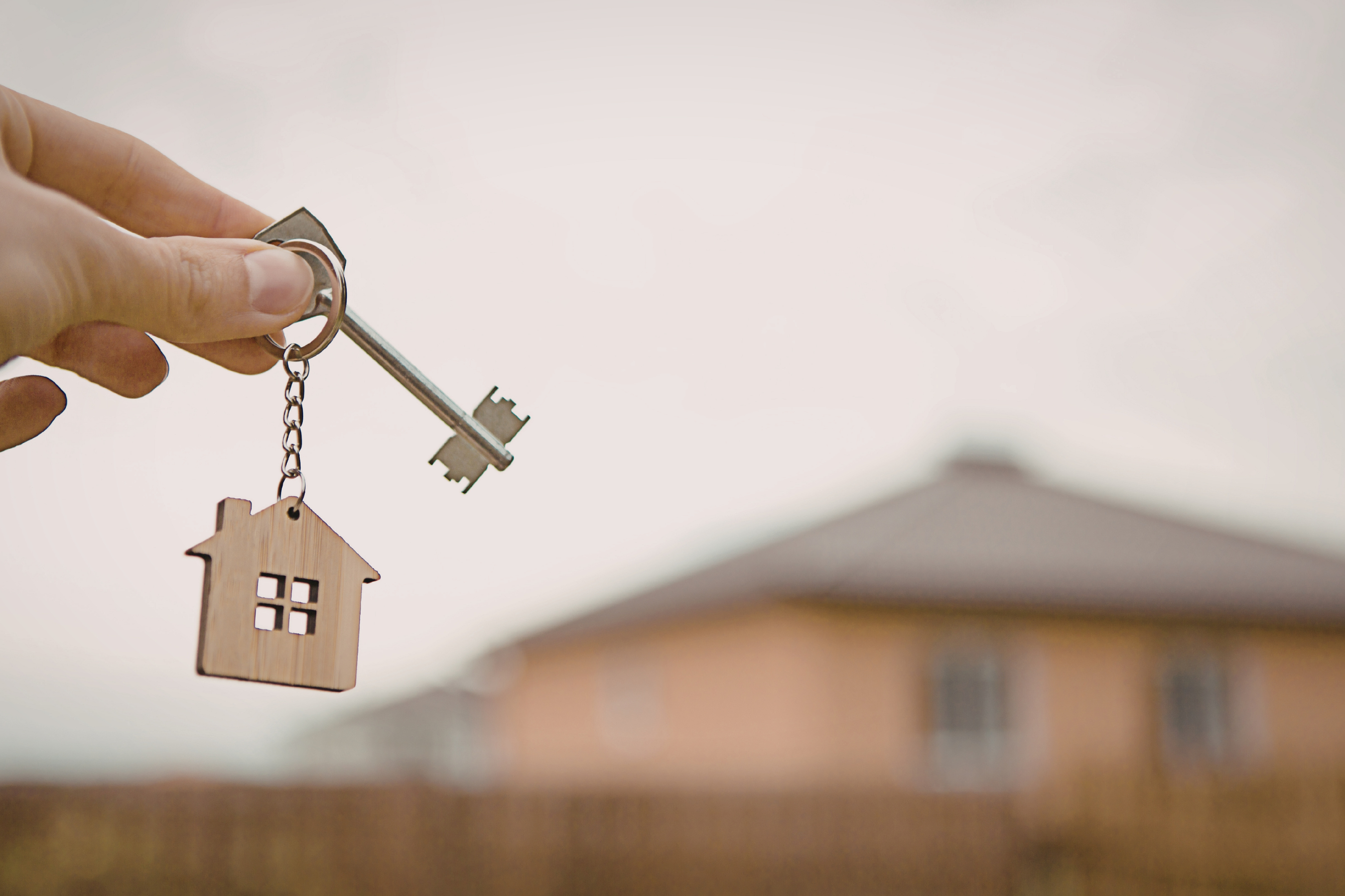 a hand holding a key with a house-shaped keychain attached with a blurry house in the background