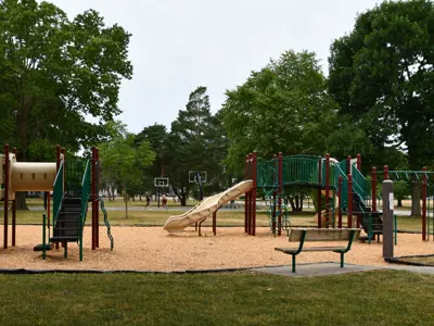 Wide view of the playground at Rev. Dr. Martin Luther King Jr. Park in Rochester, featuring multiple play structures with slides and climbing equipment surrounded by wood chips, with basketball courts, mature trees and open green space in the background.