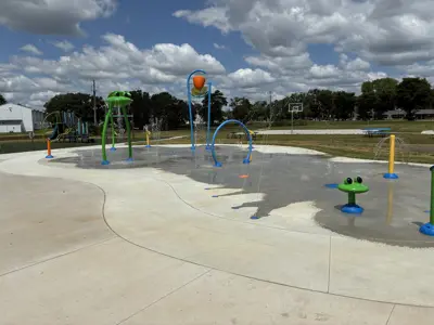 McQuillan Splash Pad at McQuillan Park featuring colorful water play elements, including spraying arches, ground jets, a tipping bucket, and frog-themed features. Water is running throughout the splash pad, with a playground and basketball court visible in the background under a partly cloudy sky.