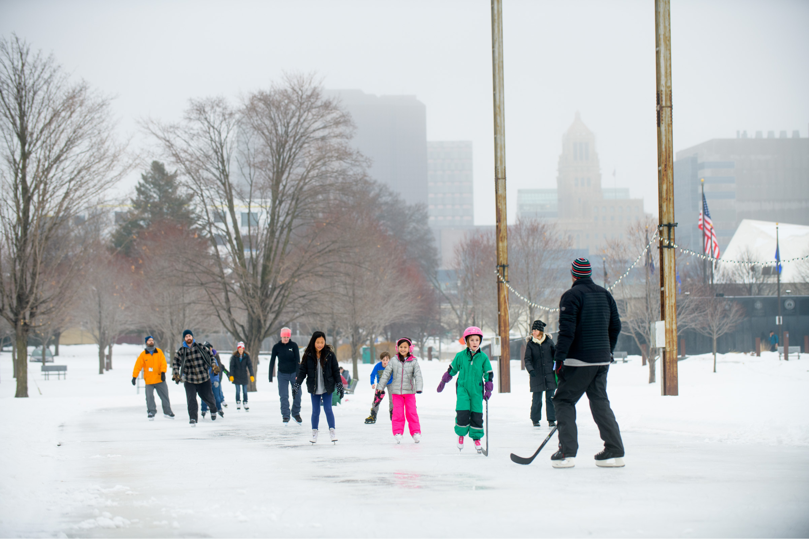 a group of people of varying ages skating on a flooded ice track with a cityscape in the background