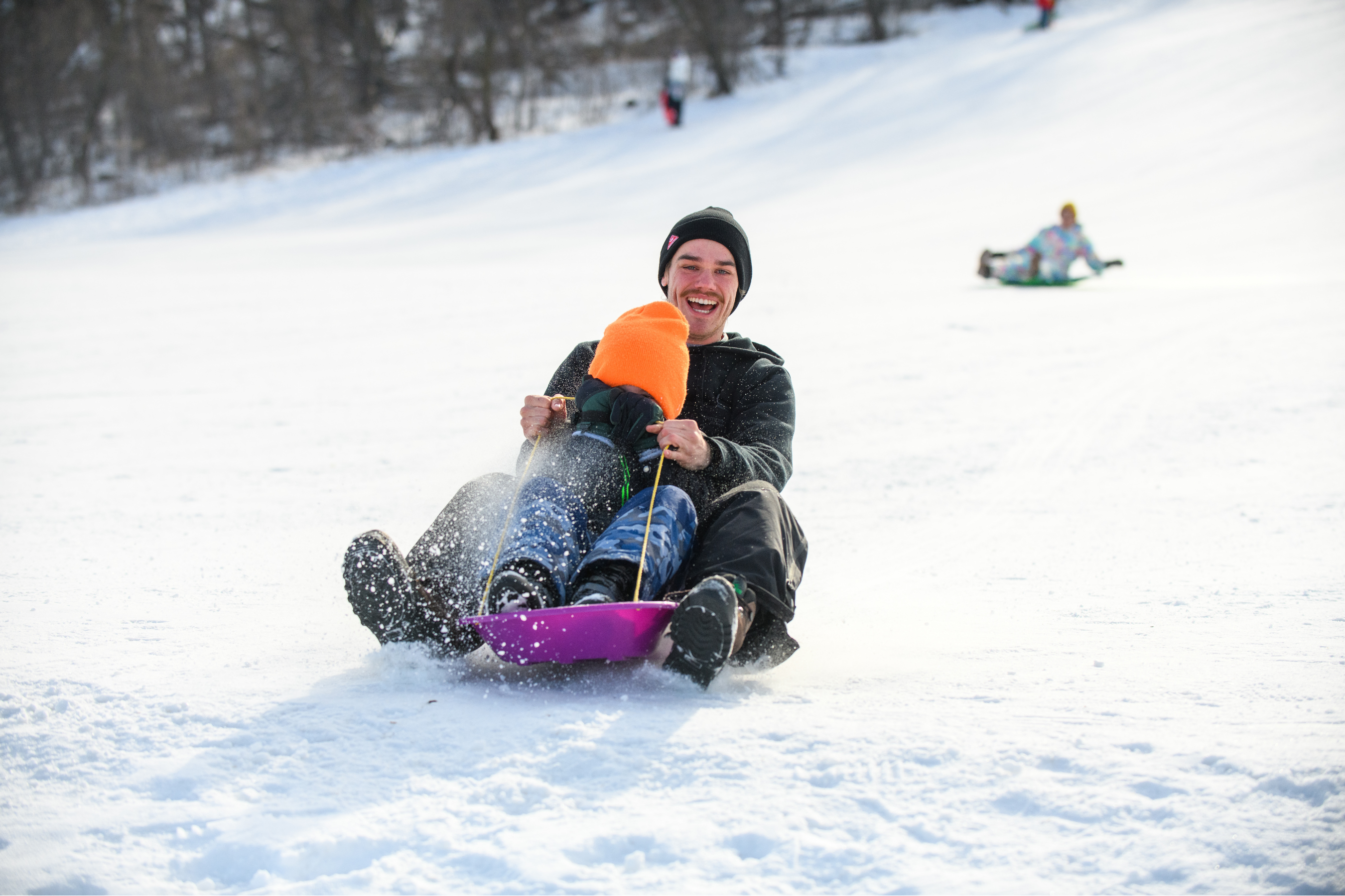 an adult smiling while sledding down a snowy hill with a child covering their face