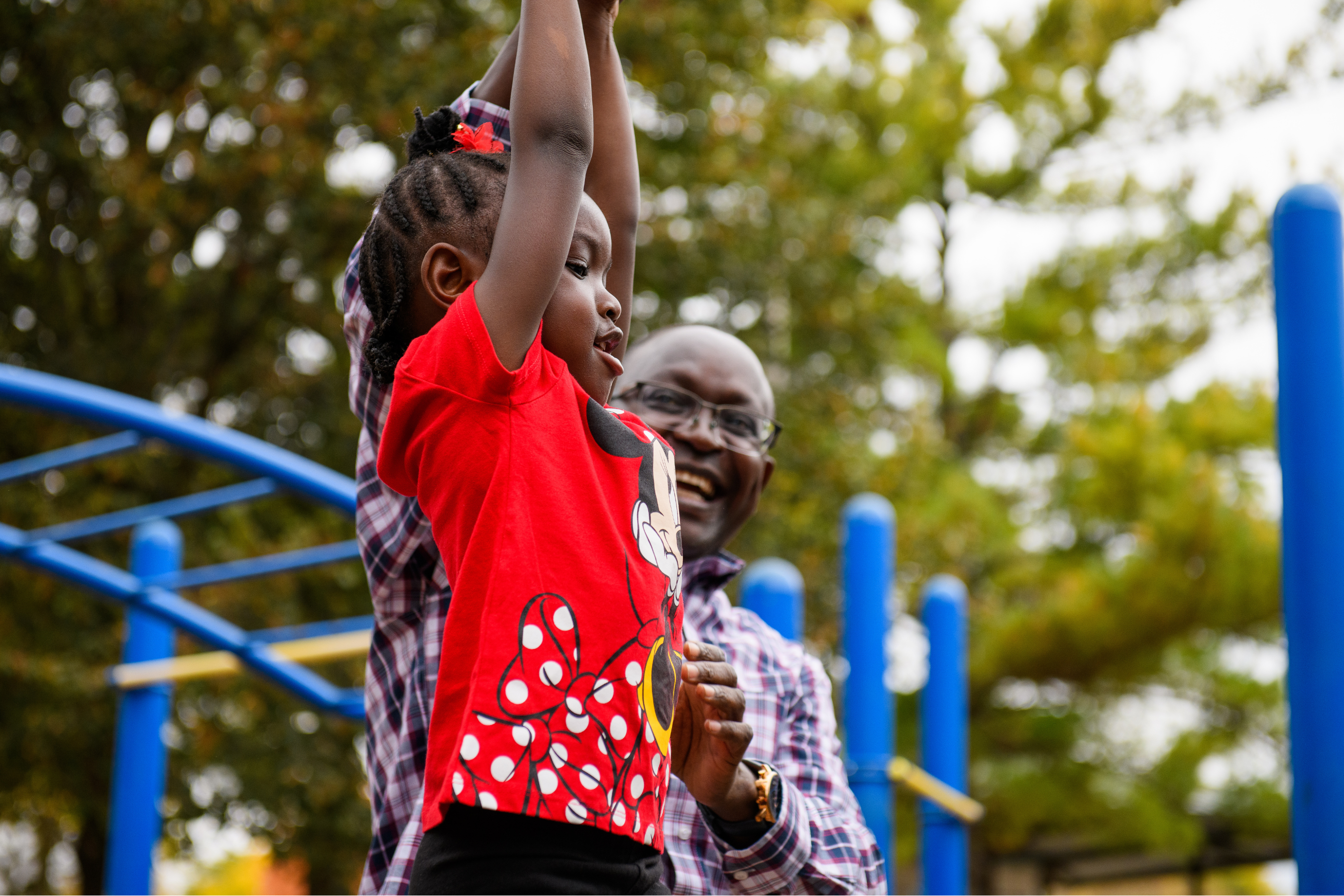 a young girl hanging from a jungle gym with an adult male standing nearby and smiling