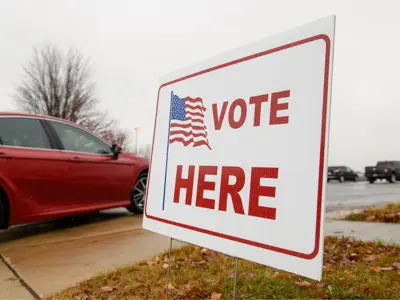 a yard sign reading 'vote here' with an american flag seen outside on a wet fall day