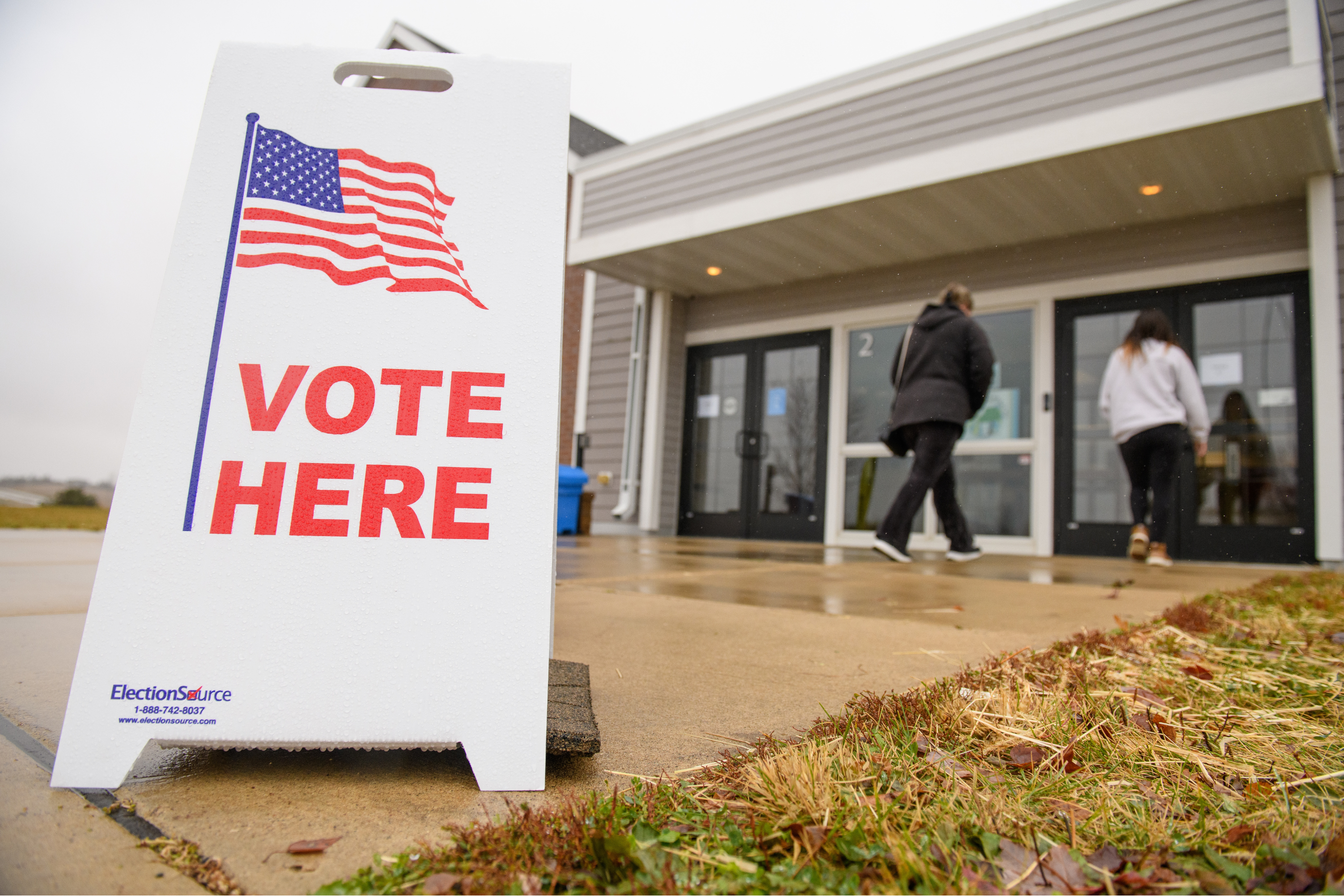 a sandwich board reading 'vote here' with an american flag with people walking into a building in the background