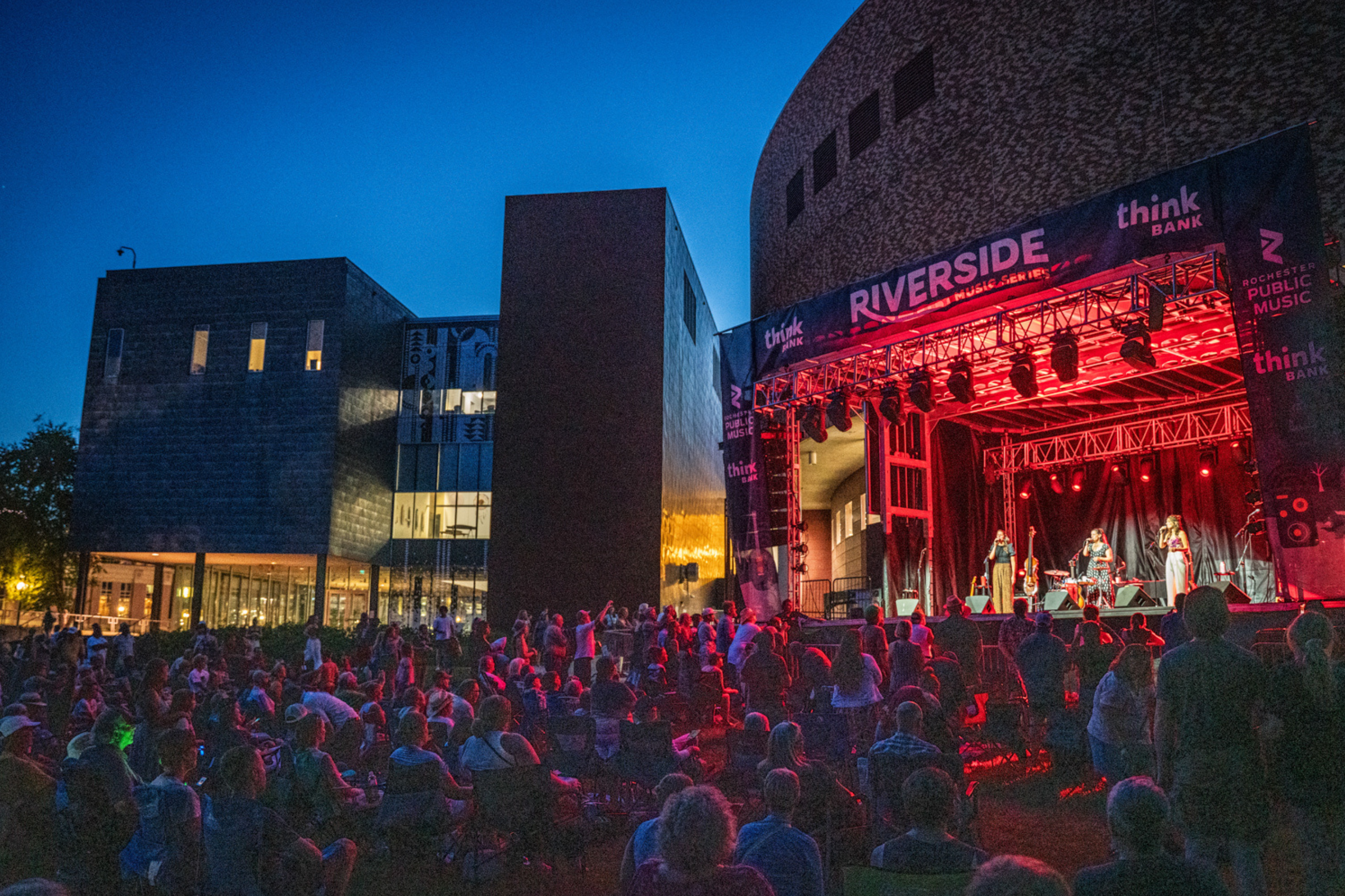 a crowd gathered in front of a stage at night with three people singing on the stage