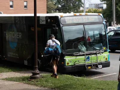 Person carrying bags boards a Rochester Public Transit bus at a curbside bus stop. The bus's route display reads "412 65th St. NW."