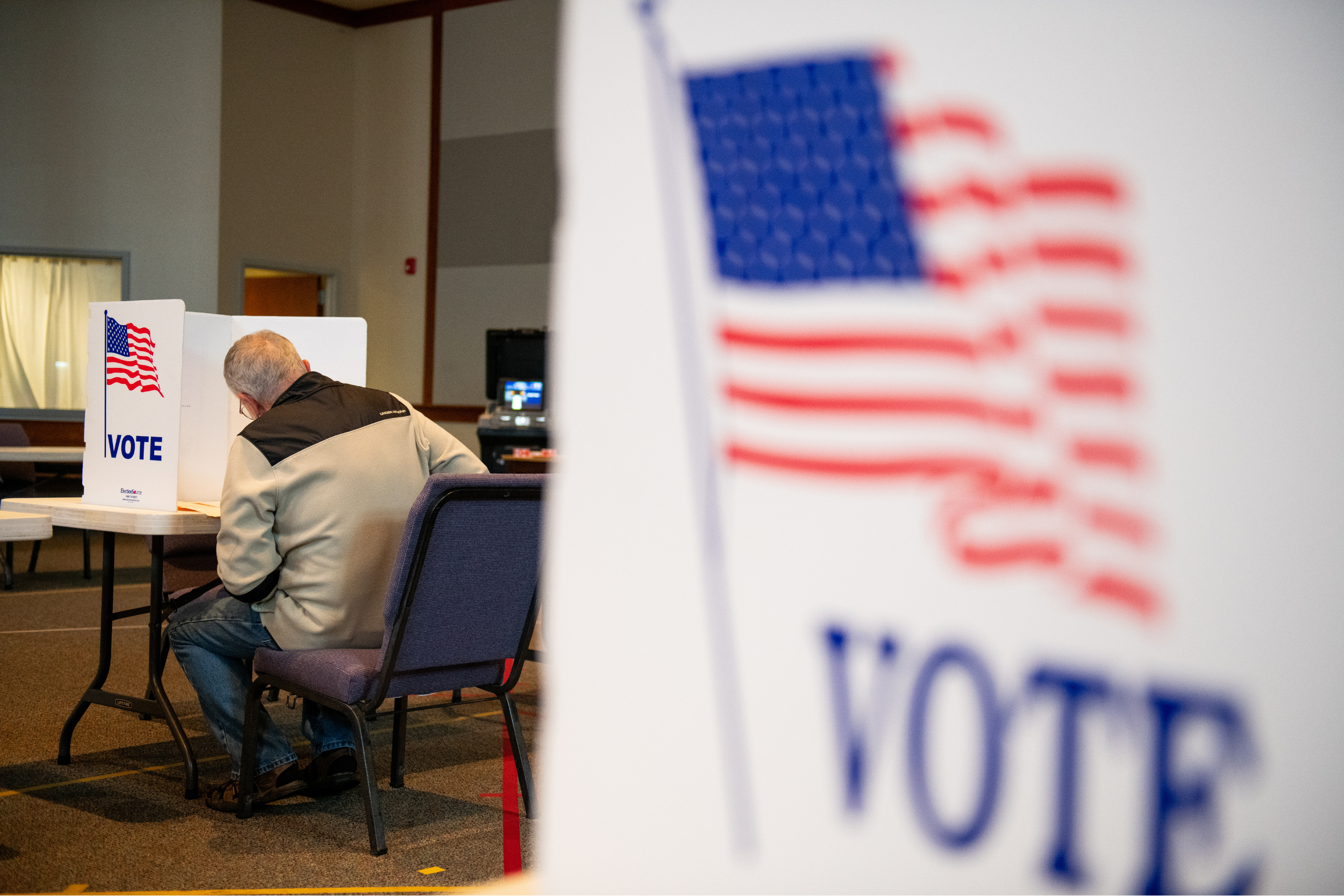 A blurred ballot box in the foreground with a man faced away filling out a ballot in the background