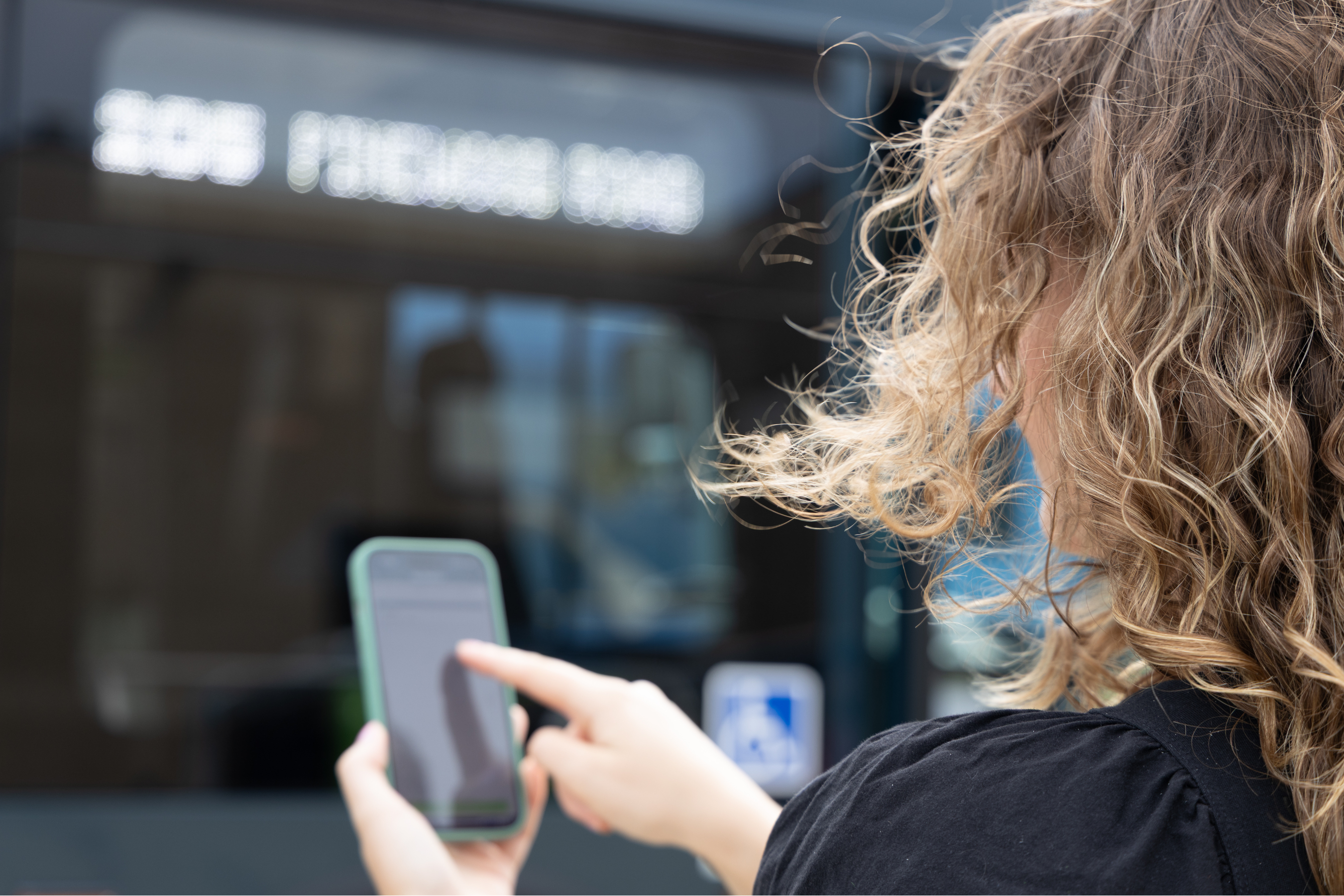 a person with curly hair facing away from the camera holding a smart phone in front of a city bus