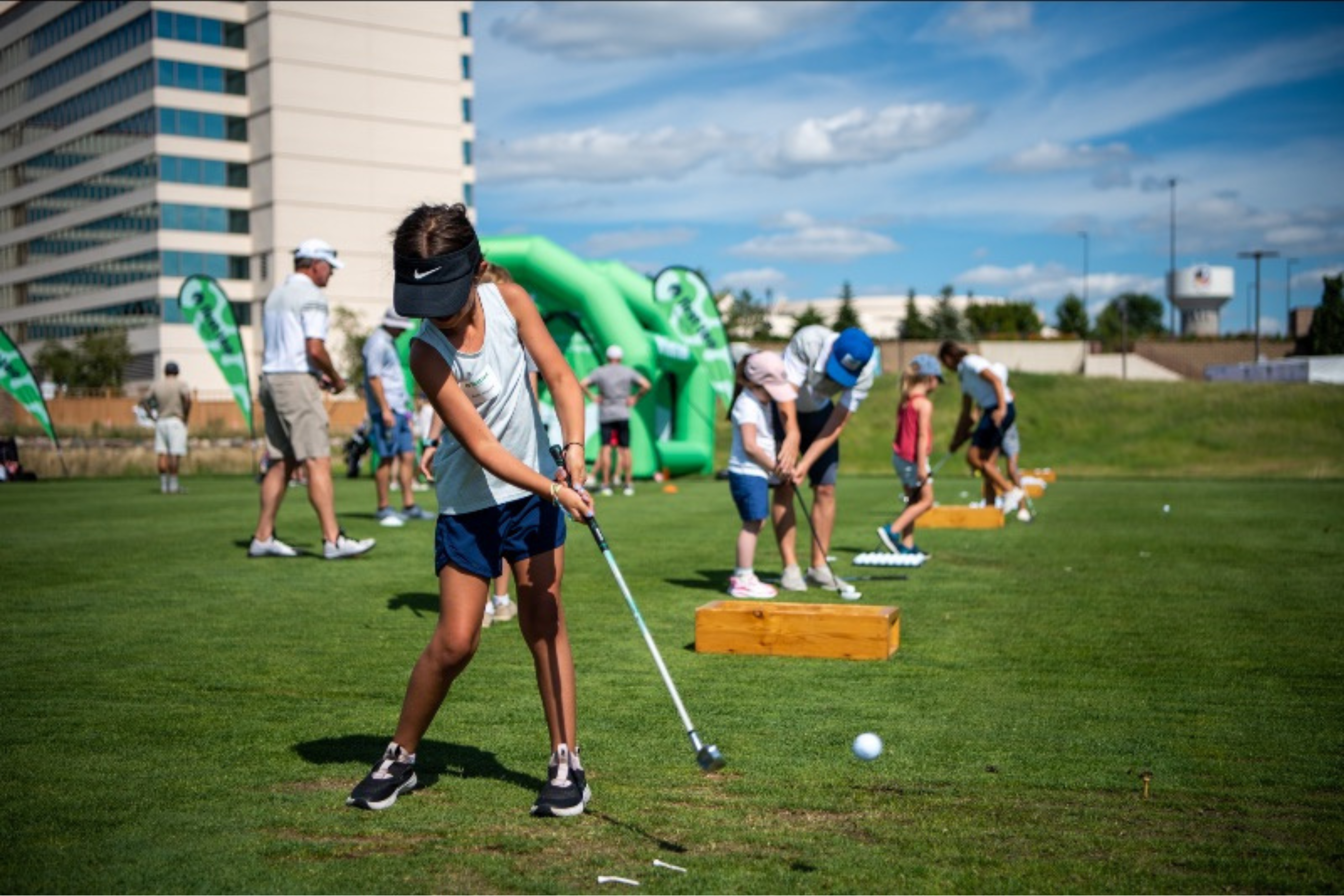 several youth golfers lined up at a driving range with various adult helpers