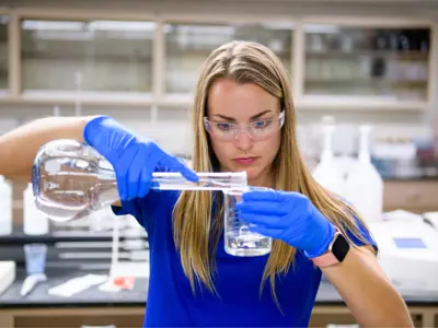 a woman pouring liquid from one beaker to another while wearing gloves and goggles in a lab setting